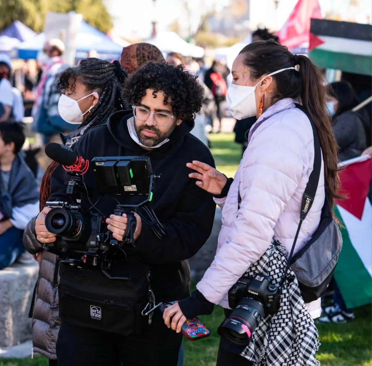 Two women and a man standing outdoors, two women wearing face masks, one of them holding a camera, the man looking at a camera or recording device, with a crowd in the background at an event or gathering.