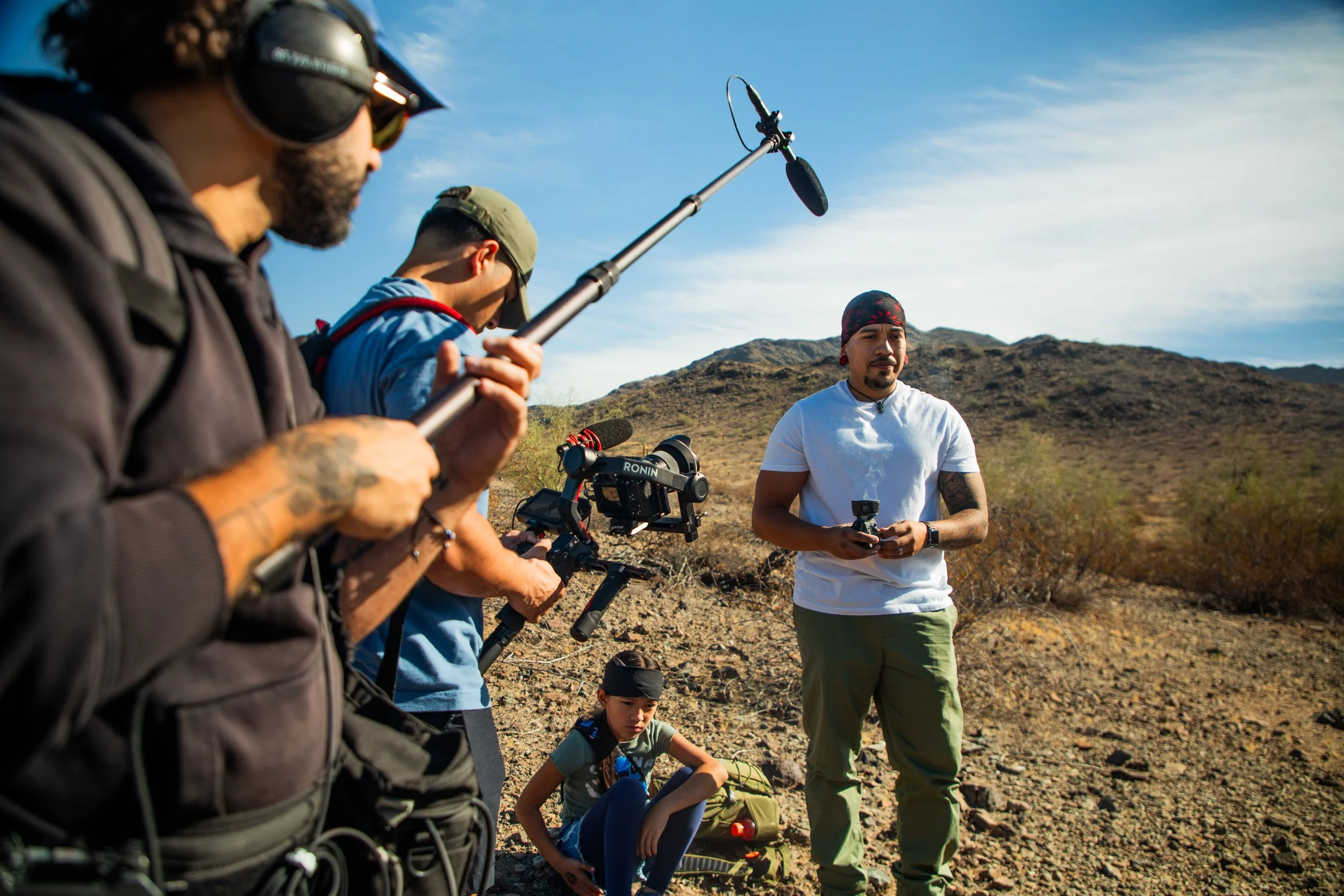 Group of four people, including a young girl, filming or taking photos outdoors in a desert landscape with mountains in the background. One man is holding a boom microphone, another has a camera mounted on a stabilizer, and a third man is holding a remote control. The girl is sitting on the ground.