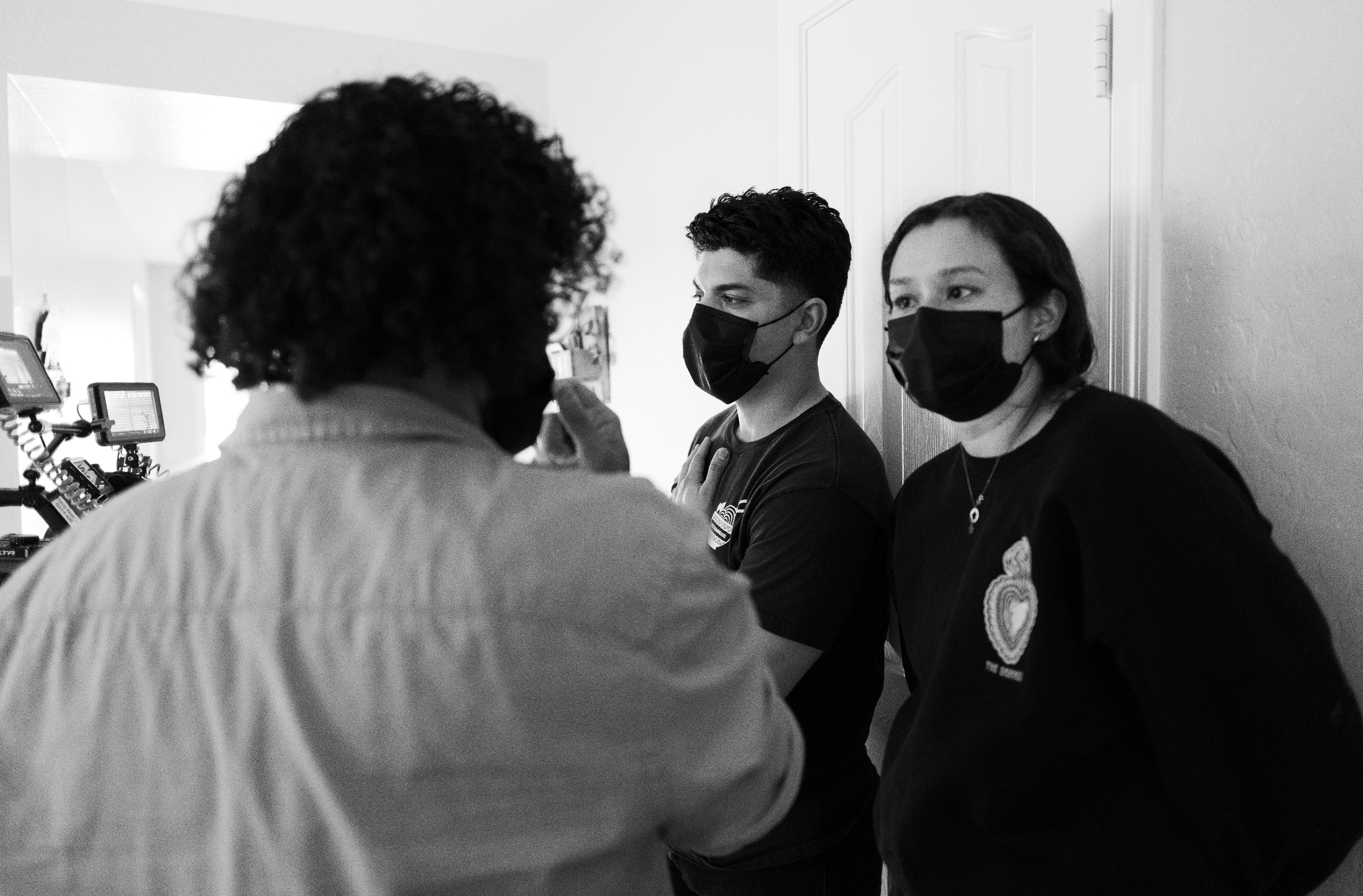 Three people wearing face masks engaged in a conversation, with one person in the foreground and two people in the background near a wall.