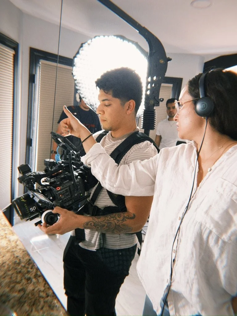 Two people working on a video shoot, one with headphones and the other with a camera, a bright ring light illuminating the scene, and several crew members in a room with windows and closed blinds.