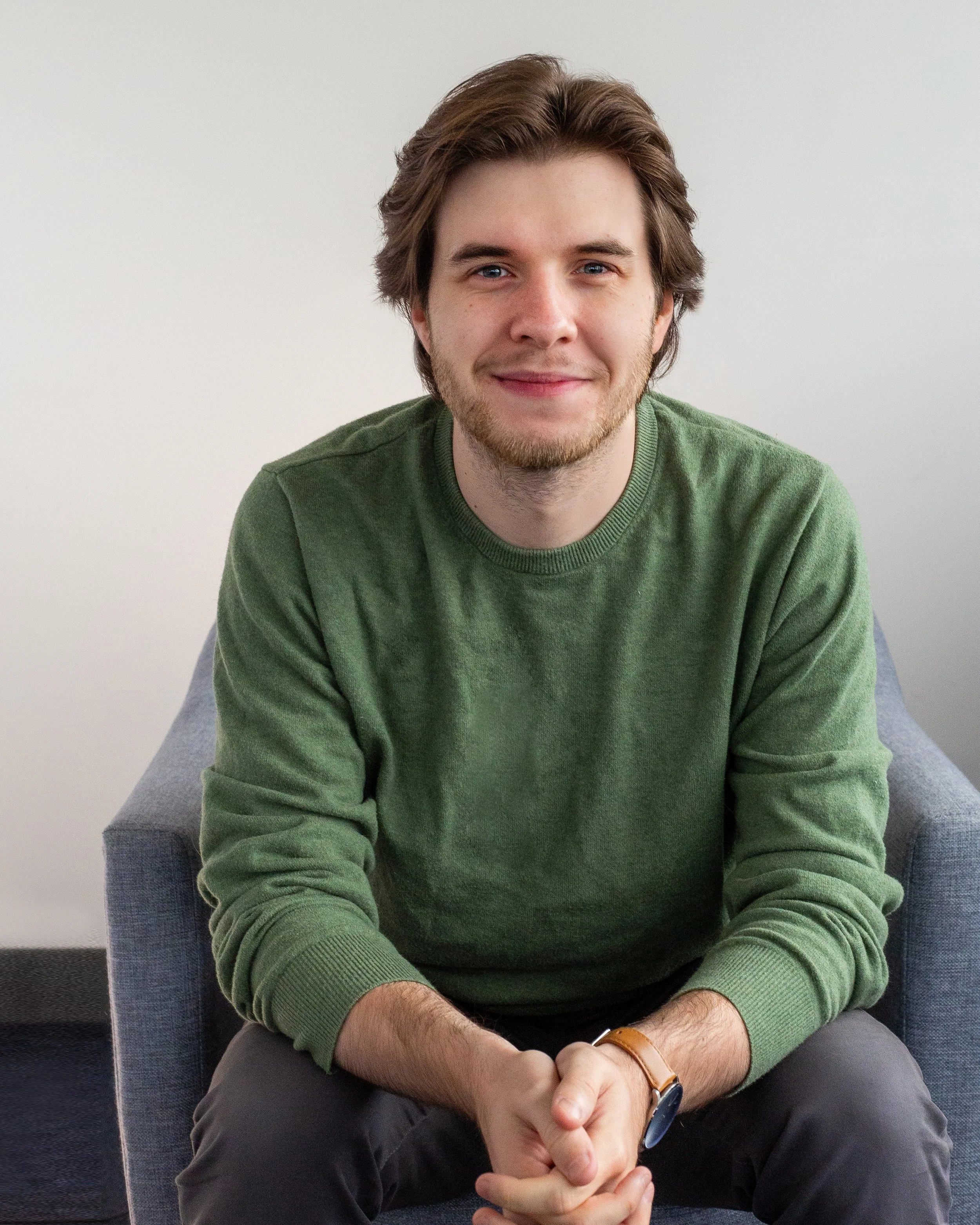 A smiling man with dark brown hair and a beard, wearing a black zip-up jacket and a white shirt, sitting indoors near large windows with a city skyline view in the background.