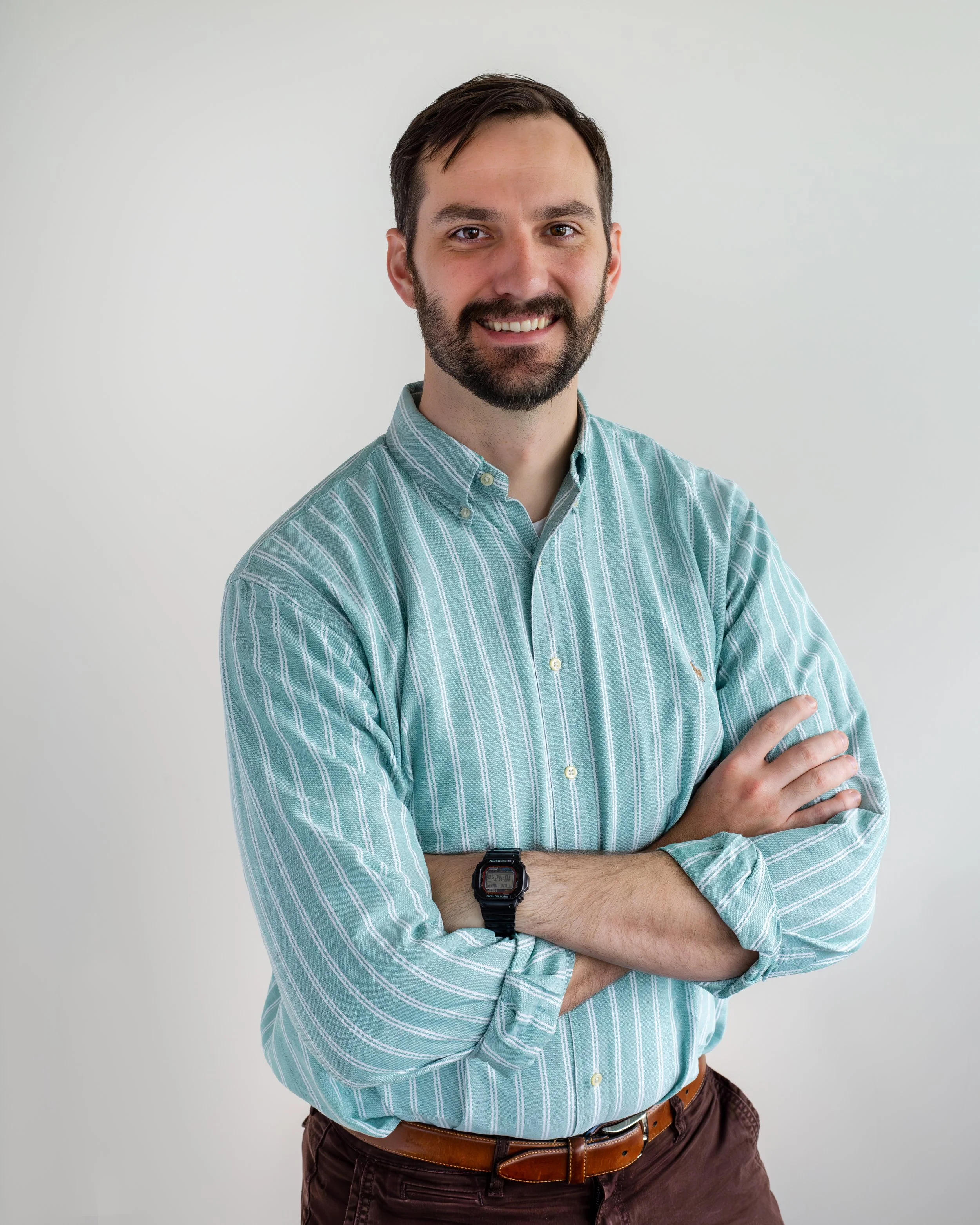 A young man with dark brown hair, a beard, and a mustache, smiling and wearing a light blue and white striped button-up shirt, sitting in front of a brick wall.