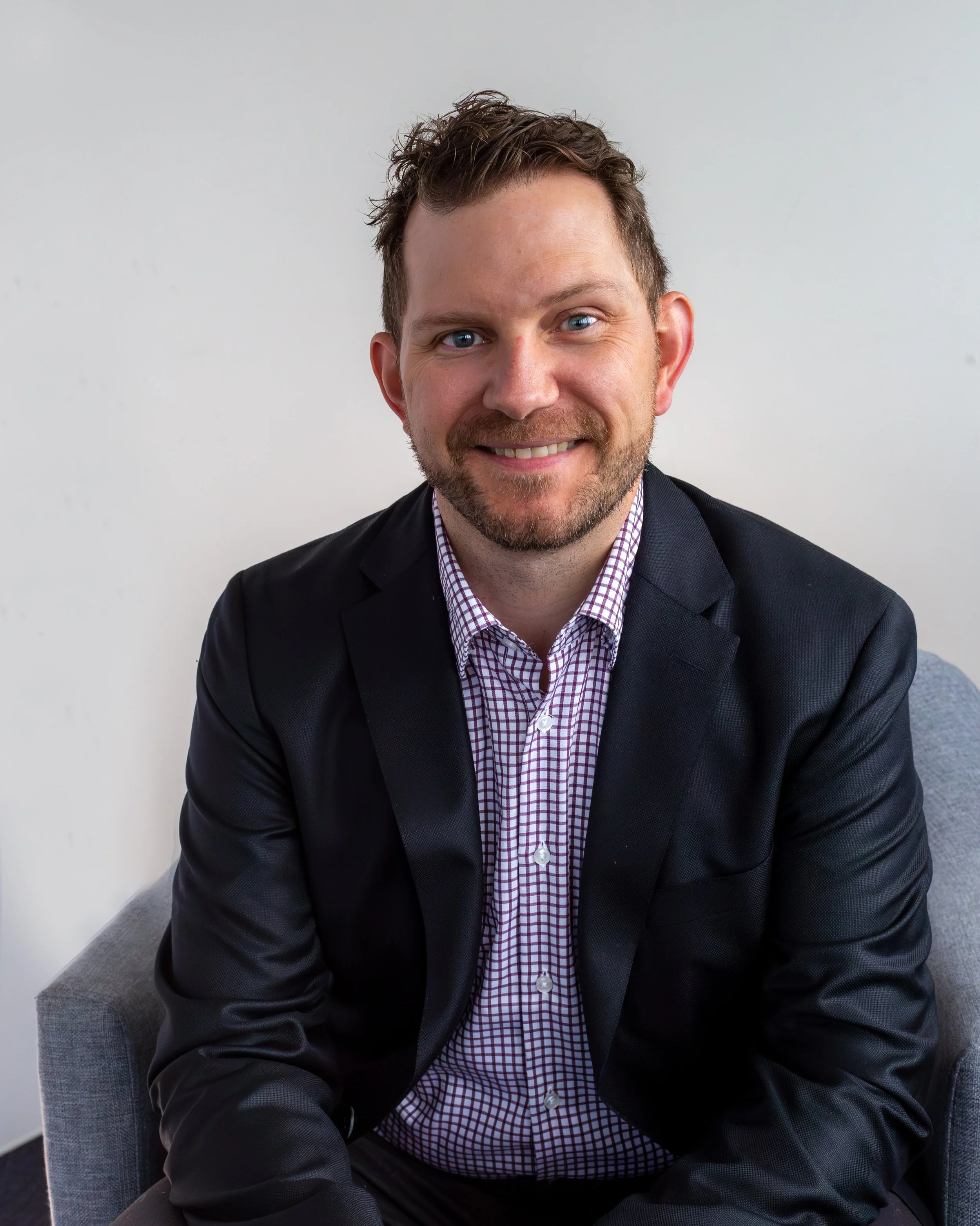 Smiling man with beard in blue checkered shirt standing near a window in a bright room.