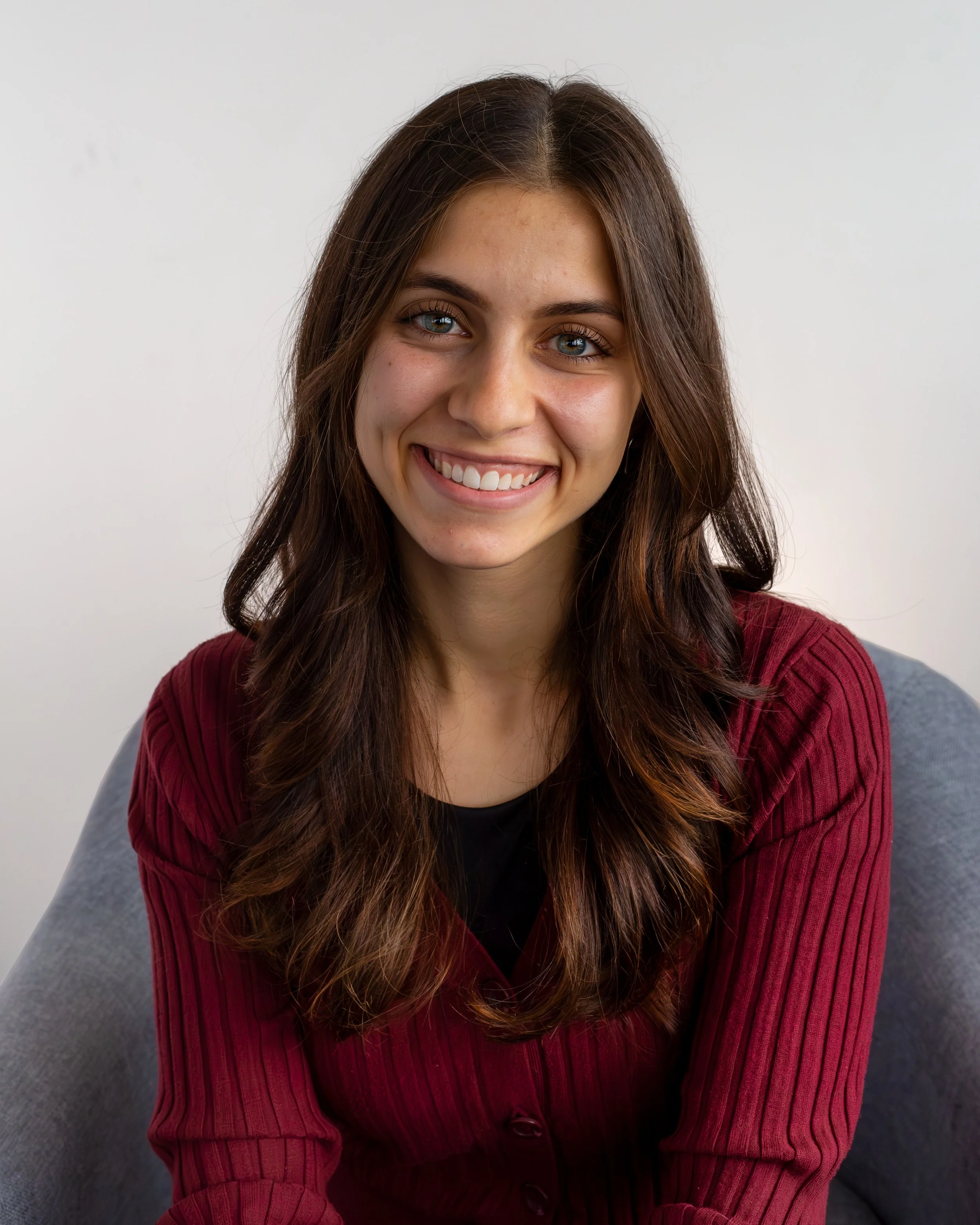 A young woman with long brunette hair and blue eyes, smiling, wearing a red sleeveless top, standing outdoors in front of a light-colored background.