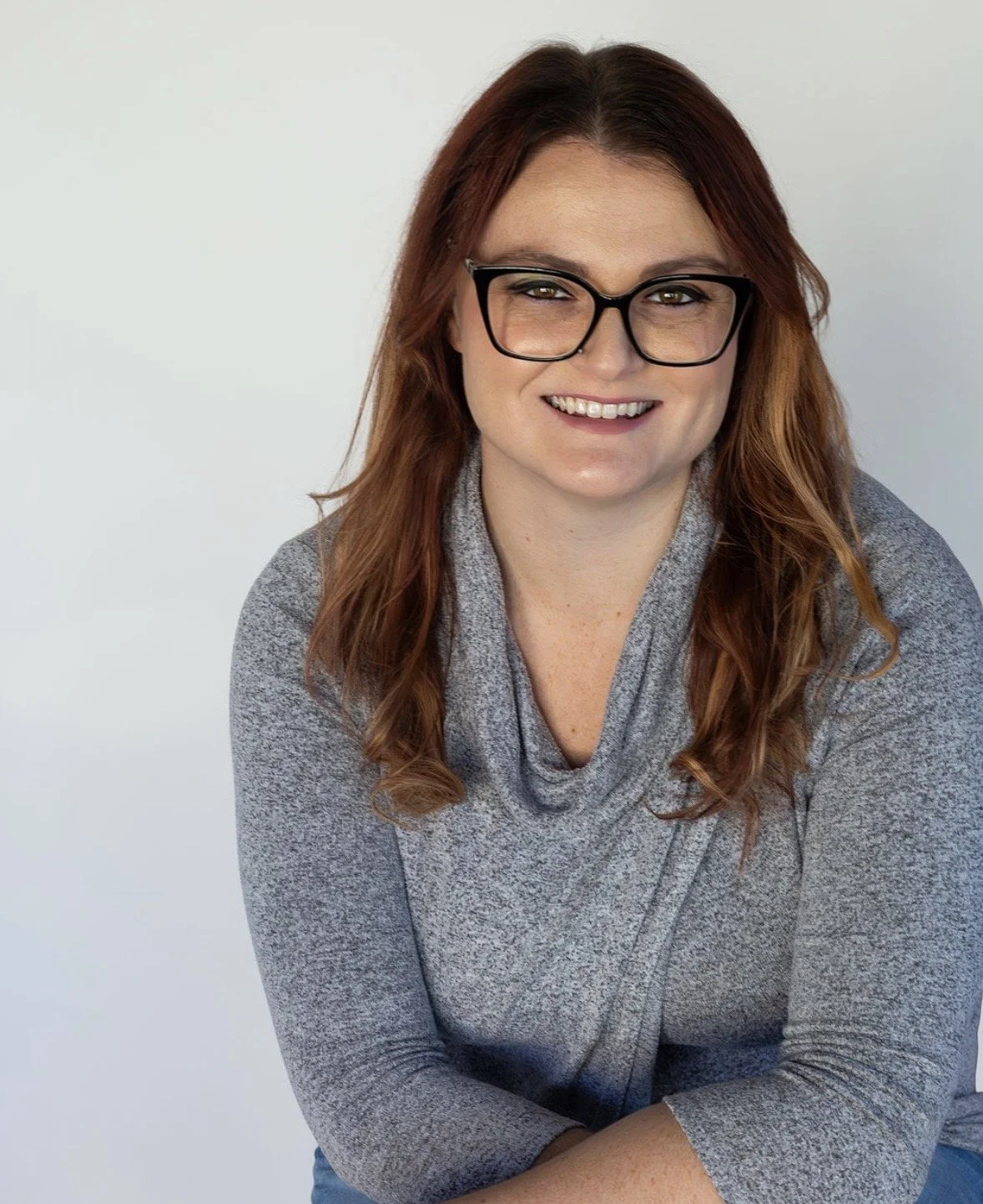 Portrait of a woman with red hair, black glasses, and a gray sweater, smiling against a plain light background.
