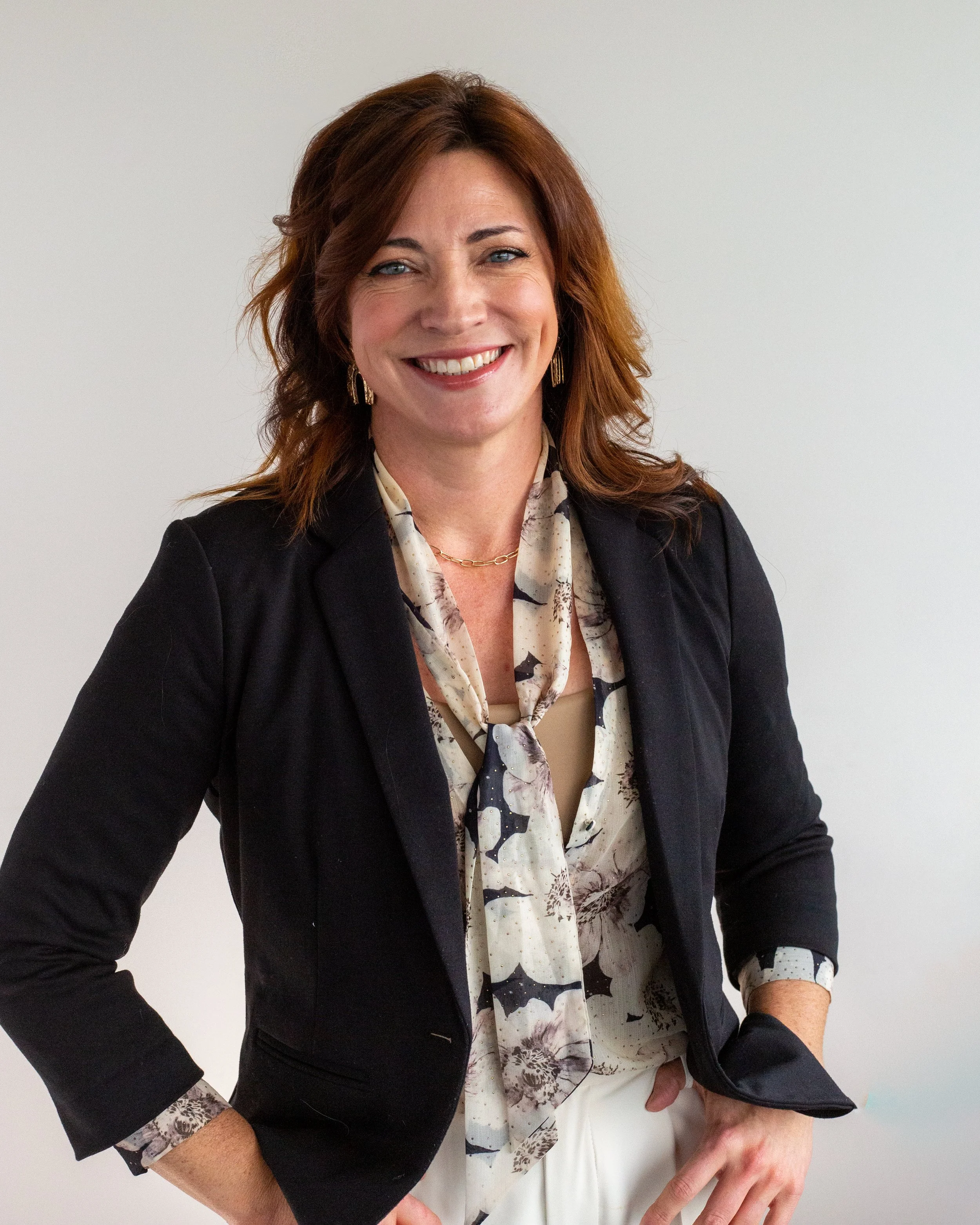 A woman with long brown hair and blue eyes smiling at the camera, standing in front of a brick wall with shelves of books.