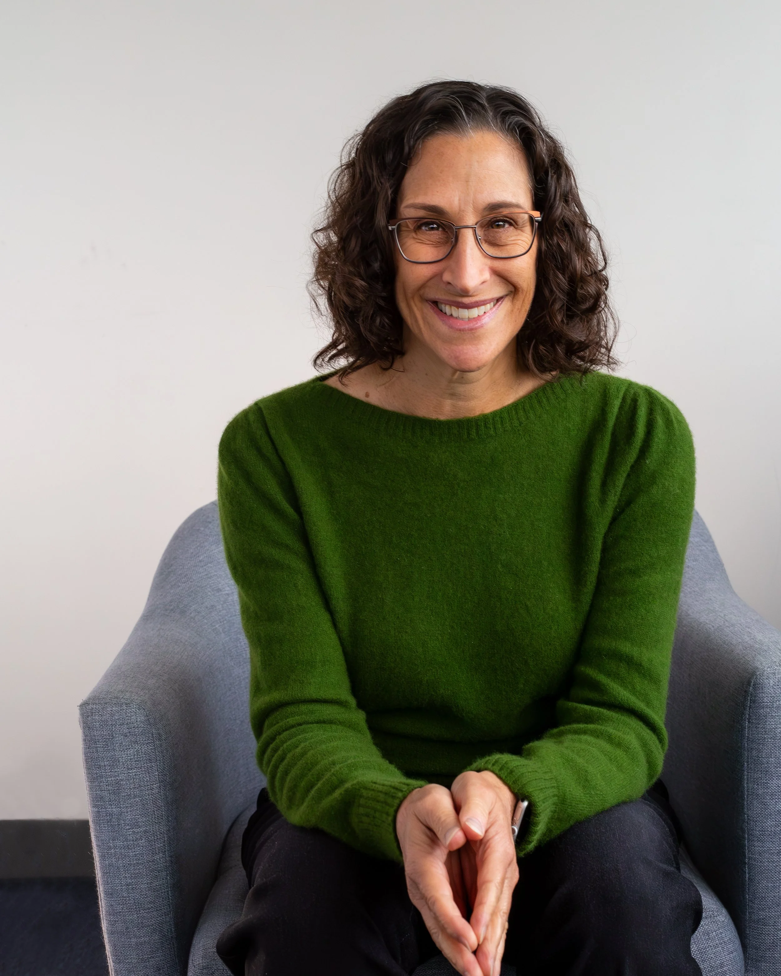 A woman with curly dark hair, glasses, and a blue sweater smiling while sitting in a room with wooden frames and bookshelves in the background.