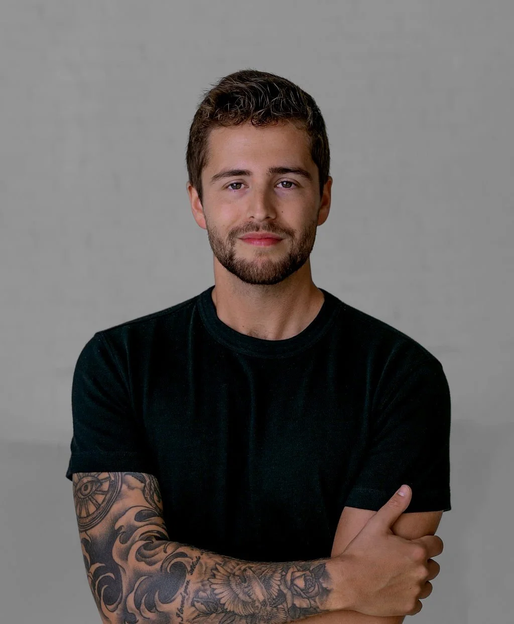 A young man with short brown hair and a beard, wearing a black t-shirt, standing with arms crossed against a plain gray background.