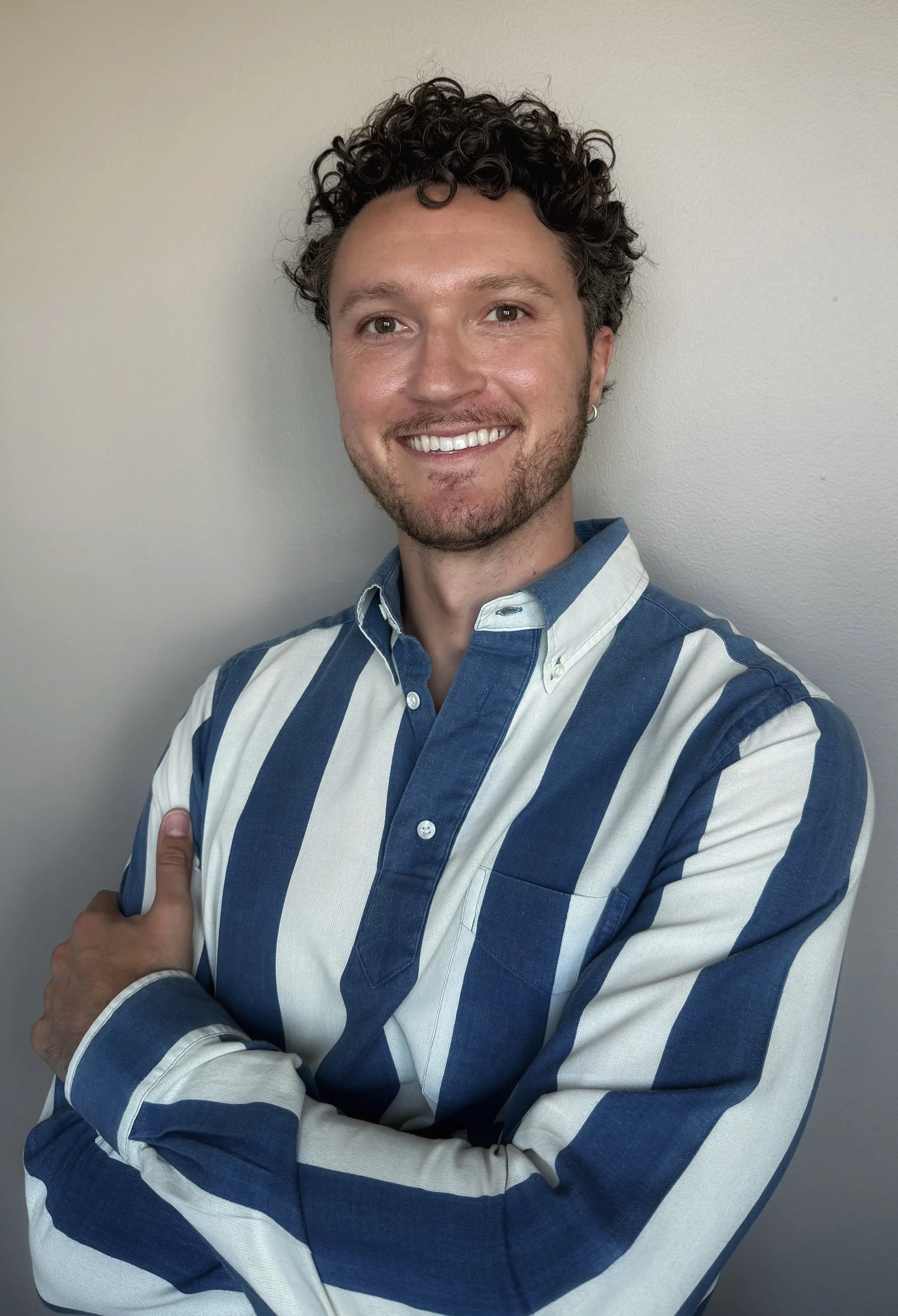 A smiling man with curly dark hair, wearing a blue and white striped button-up shirt, standing against a plain wall with his arms crossed.