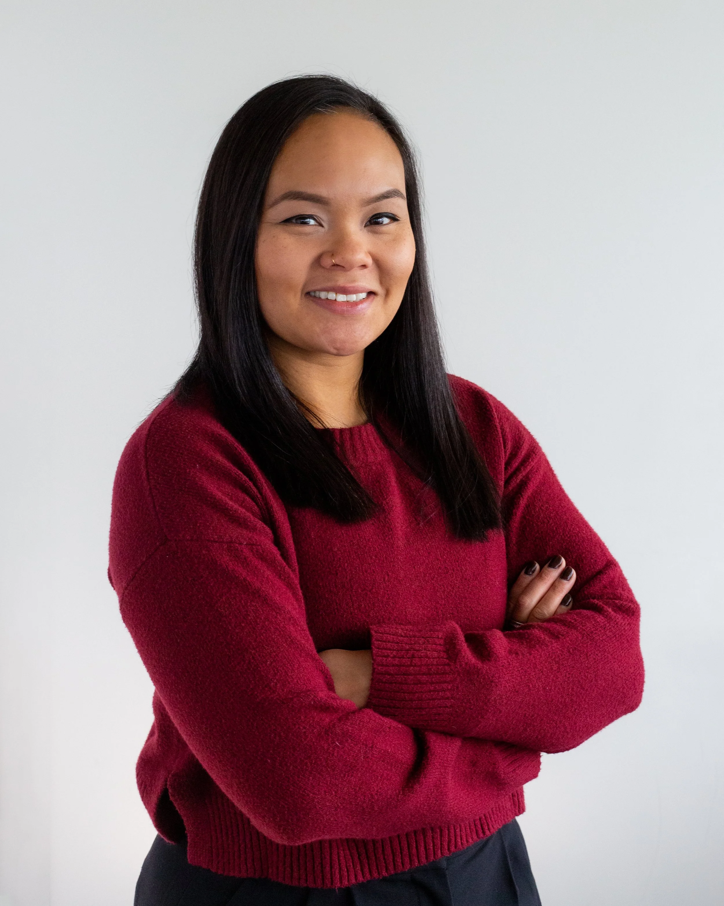 Portrait of a smiling woman with long black hair, wearing a dark shirt and an olive green jacket, standing against a plain white background.