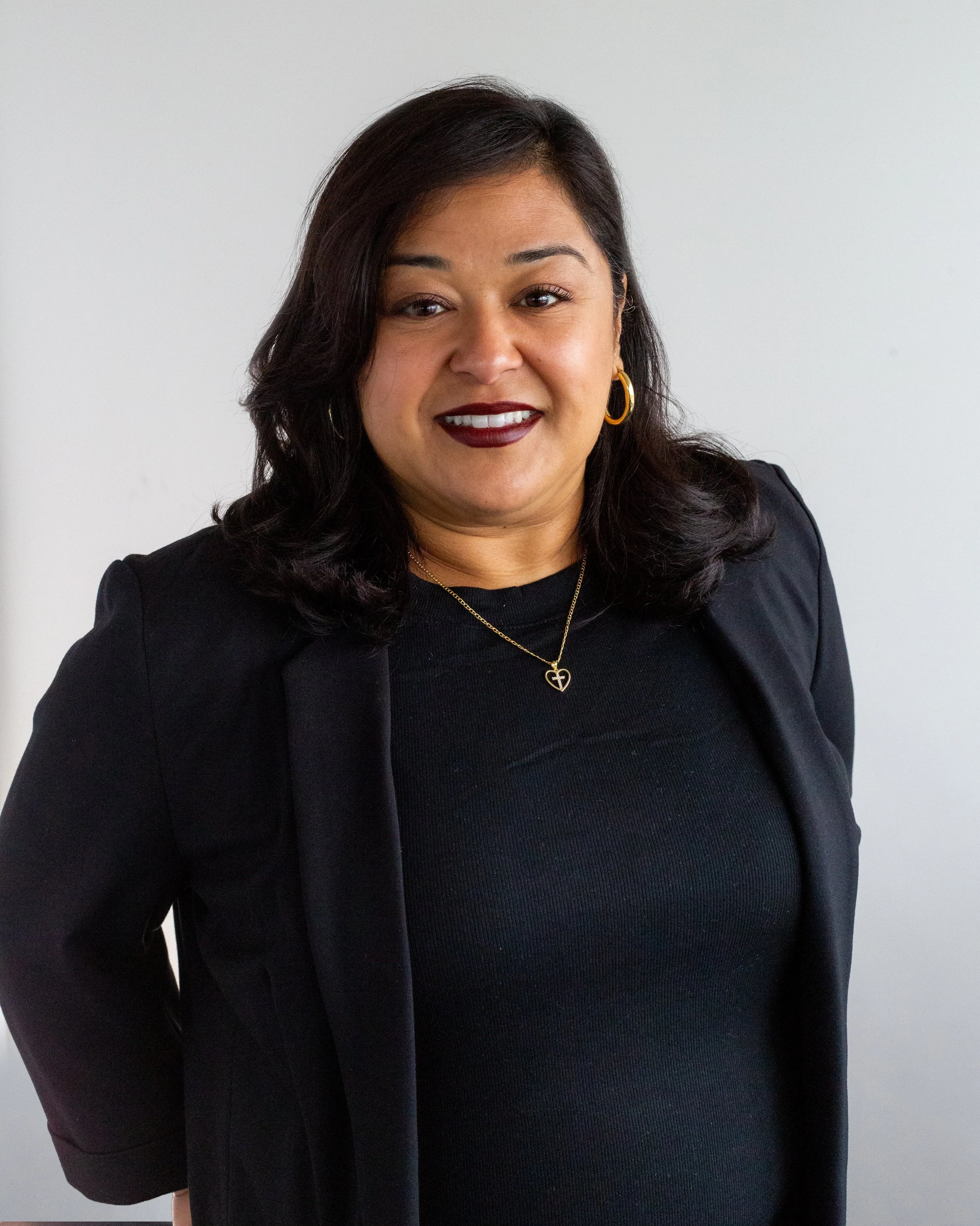 A woman with shoulder-length dark hair smiling at the camera, wearing a black top and a silver necklace with a circular pendant, standing against a light-colored wall.