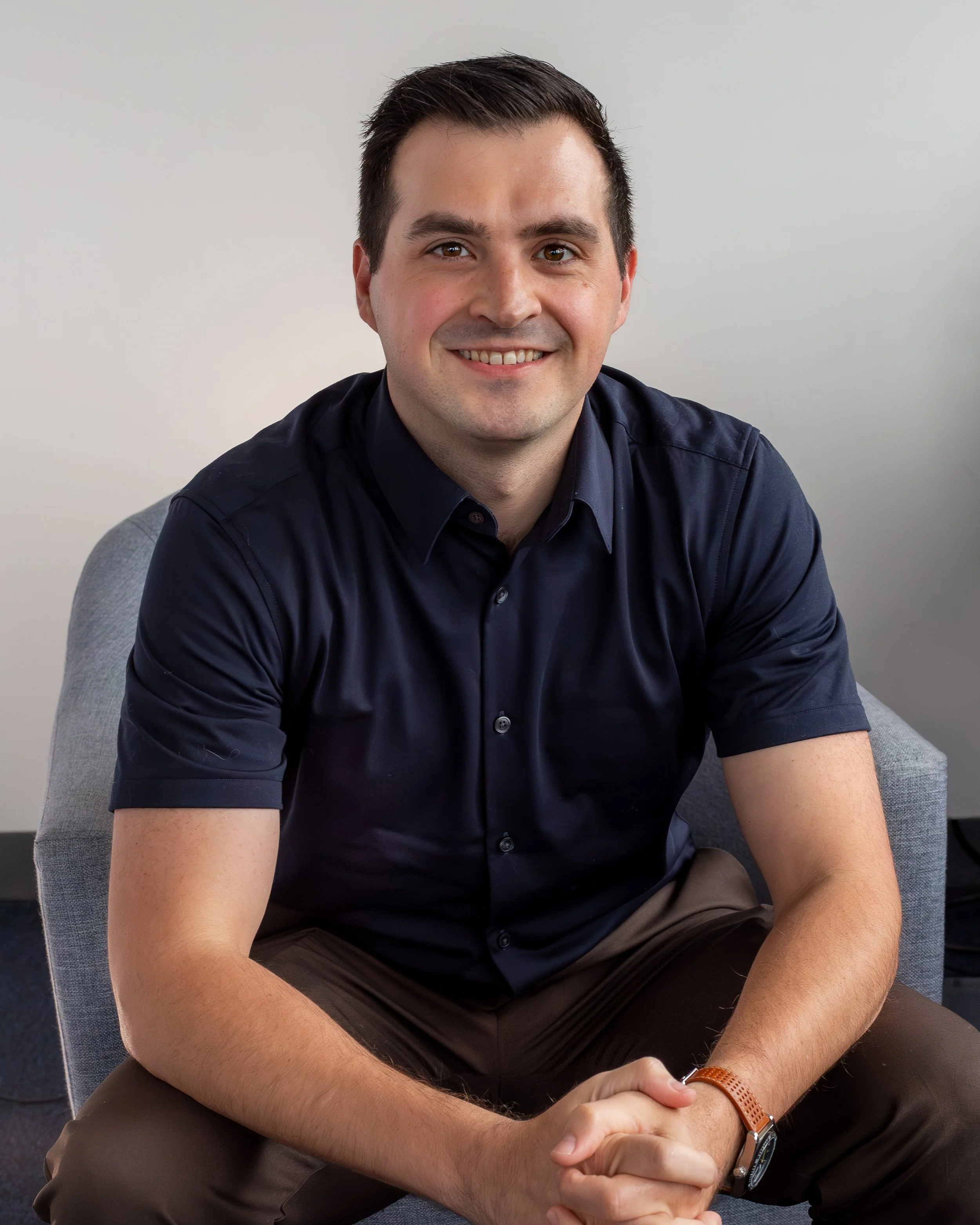 A man in a light gray blazer and white shirt smiling outdoors next to a wooden fence with green trees in the background.