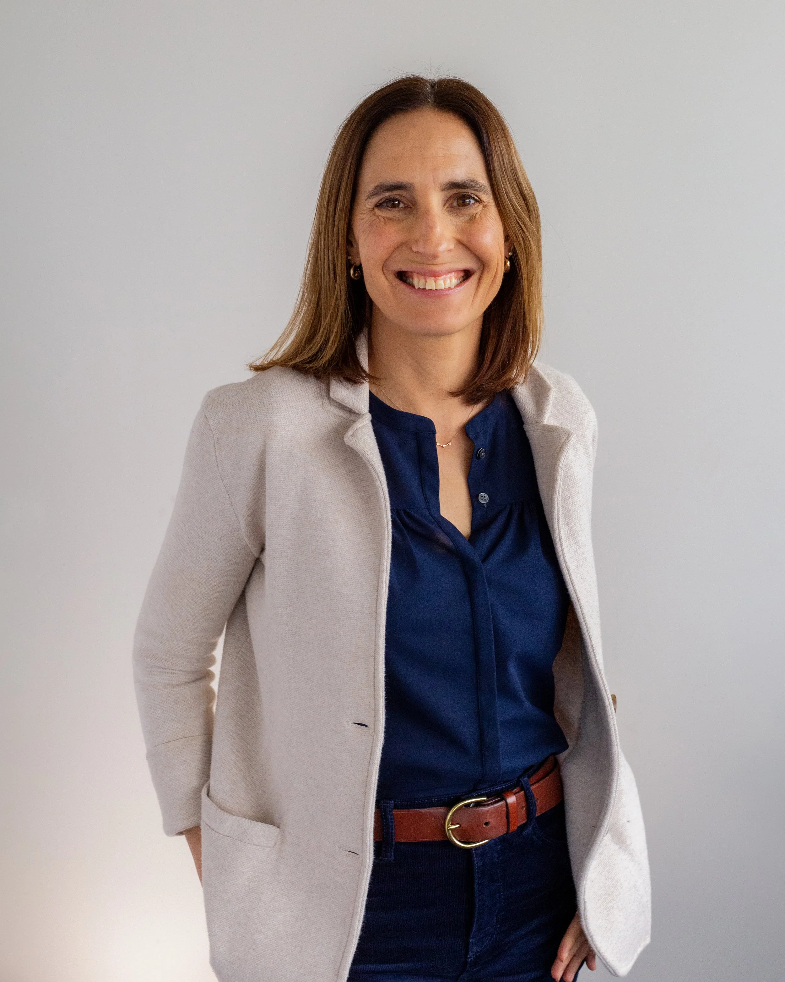 A woman with shoulder-length brown hair wearing a brown sweater with leather accents and blue jeans, standing indoors against a white brick wall, smiling at the camera.