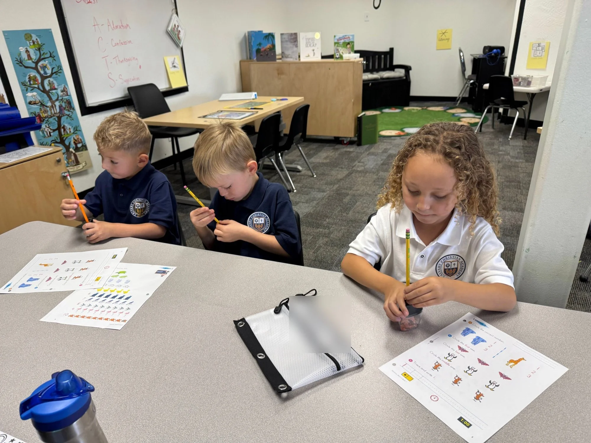 Three Kindergarten students at Calvary Christian Academy work on their math worksheets at a classroom table.