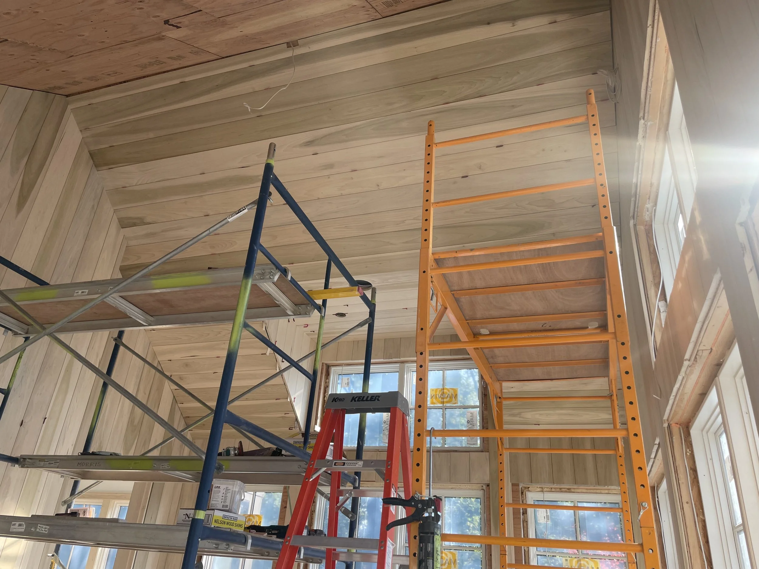 Interior of a room under construction, with wooden wall paneling and scaffolding. There is a ladder and a window with sunlight shining through.