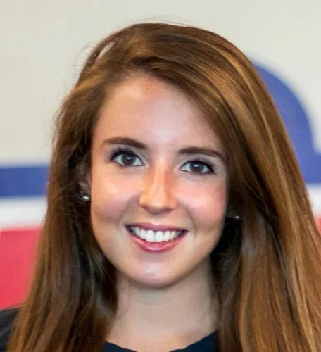 A young woman with long, reddish-brown hair smiling at the camera, with a blurry background featuring a blue and red design.
