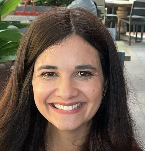 Close-up of a woman smiling with long brown hair in an outdoor cafe setting.