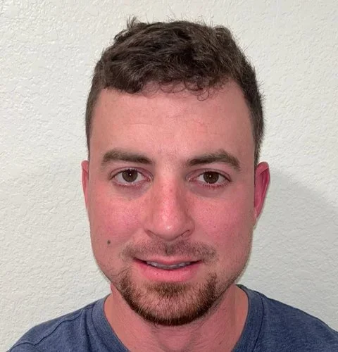 Headshot of a young man with short brown hair, light complexion, and a goatee, wearing a blue shirt, standing against a plain light-colored wall.