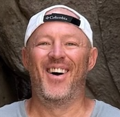 A smiling man with a beard, wearing a white cap backward, outdoors against a rock background.