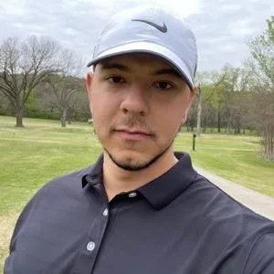Young man wearing a white Nike cap and black polo shirt standing outdoors on a golf course with trees and grass in the background.