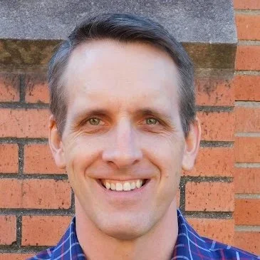 A man with short brown hair and blue eyes smiling in front of a brick wall.