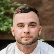 Close-up of a young man with short dark hair and a trimmed beard, wearing a white shirt, outdoors with a blurred natural background.