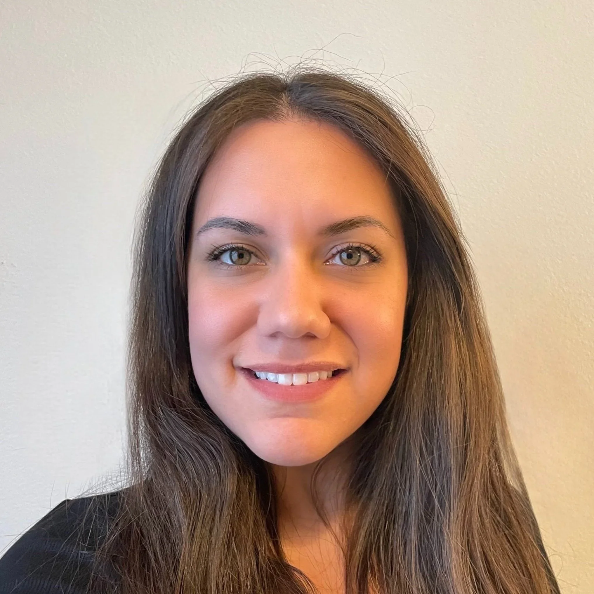 Close-up of a smiling woman with long brown hair, green eyes, and light skin, wearing a black top, against a plain beige wall background.