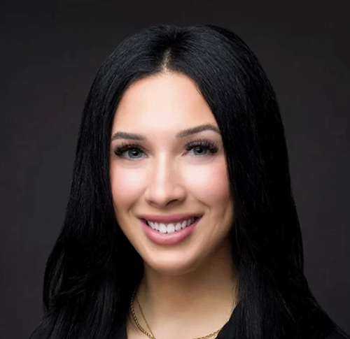 Close-up portrait of a young woman with long black hair, fair skin, and a friendly smile, against a dark background.