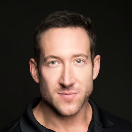 Portrait of a man with short dark hair, light eyes, and slight stubble, wearing a black shirt, against a dark background.
