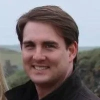 A young man outdoors with a background of green grass and sky, smiling at the camera.