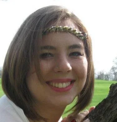 Close-up of a smiling young woman with shoulder-length hair outdoors, with a tree and grassy field in the background.