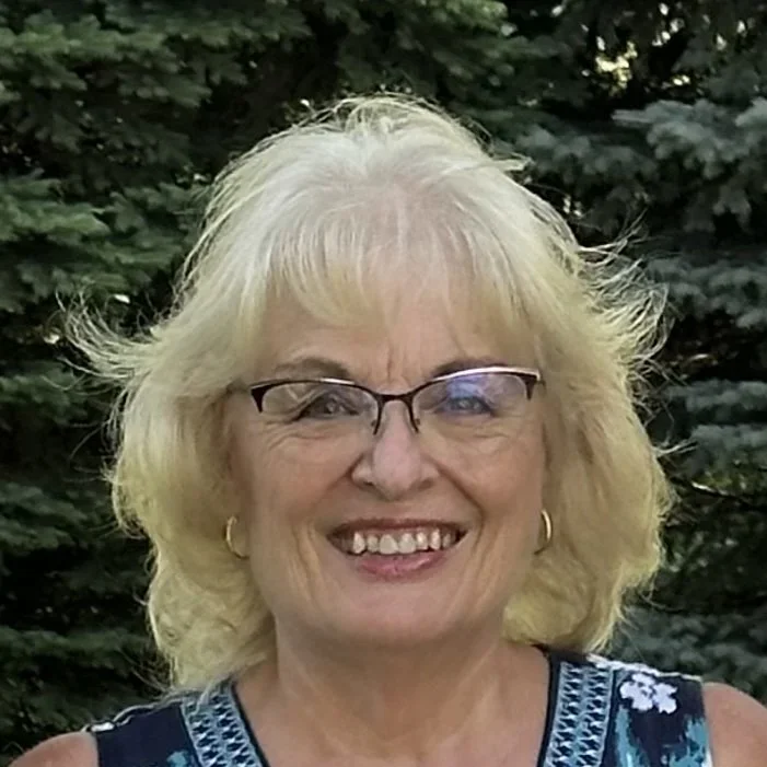 A smiling woman with blonde hair and glasses, wearing a sleeveless blue patterned top, standing outdoors in front of green trees.