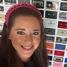 A smiling woman with long brown hair wearing a pink headband, standing in front of a wall with vintage audio cassette tapes.