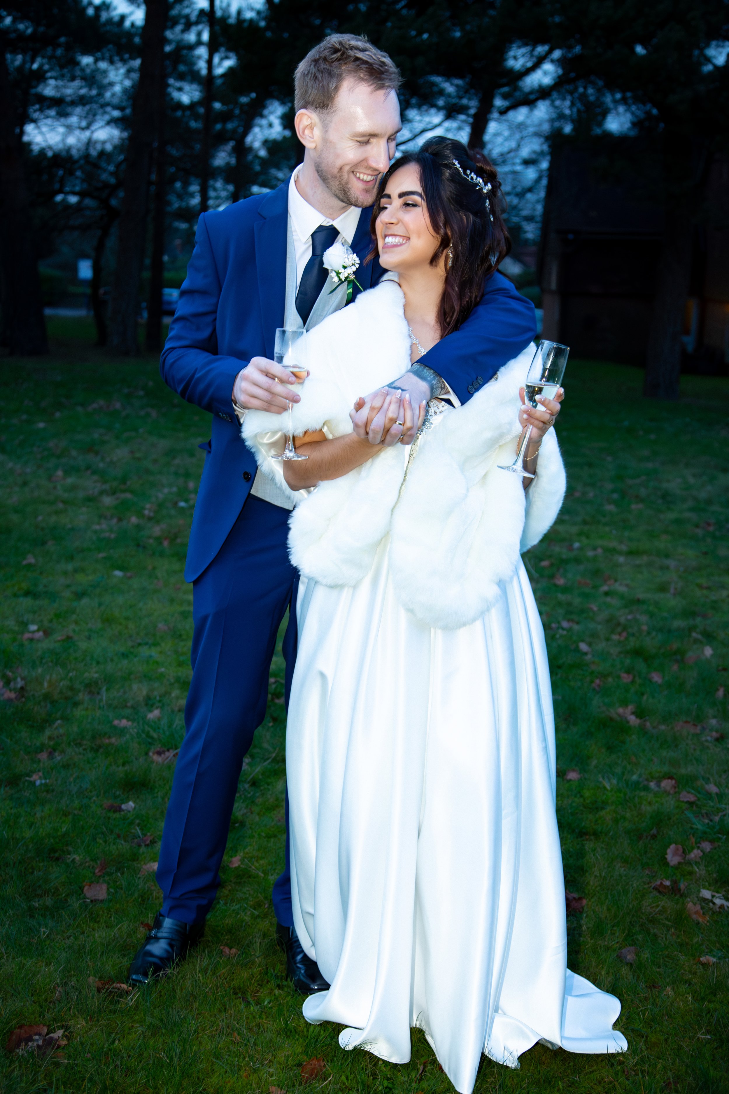 Bride and groom embrace outdoors, holding champagne glasses. The bride wears a white dress and fur shawl, while the groom is in a blue suit.