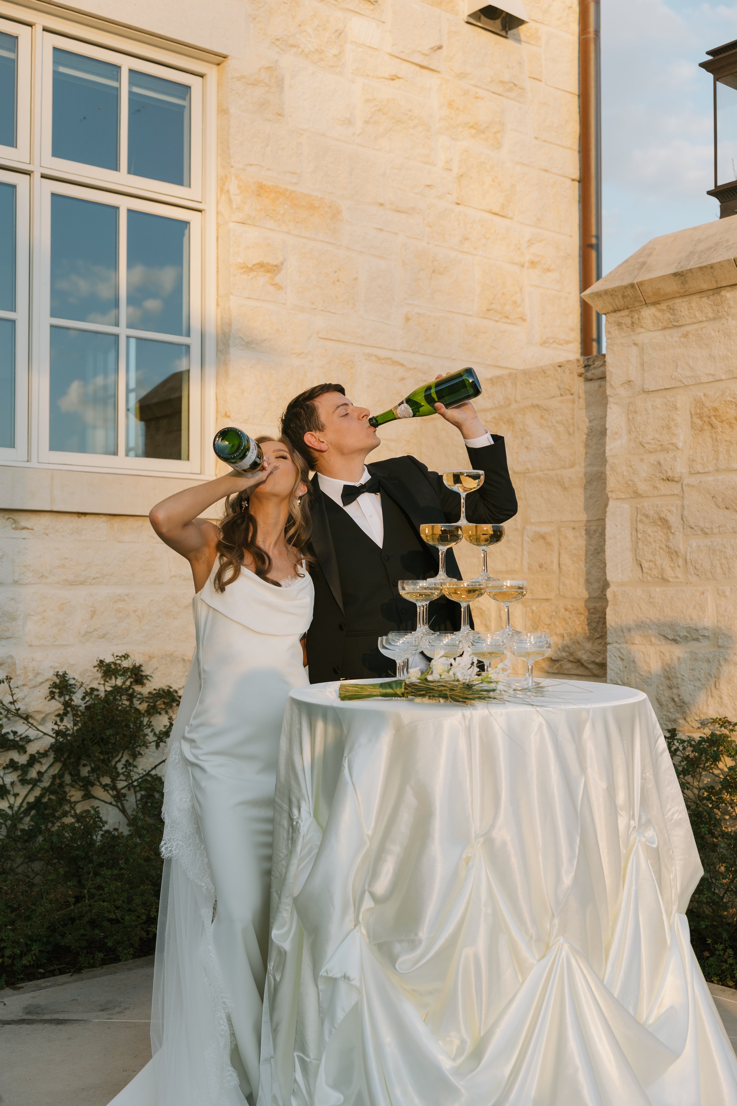 A bride and groom in formal attire standing outside near a stone building with a window, toasting with champagne bottles over a tower of empty champagne glasses on a table, during sunset.