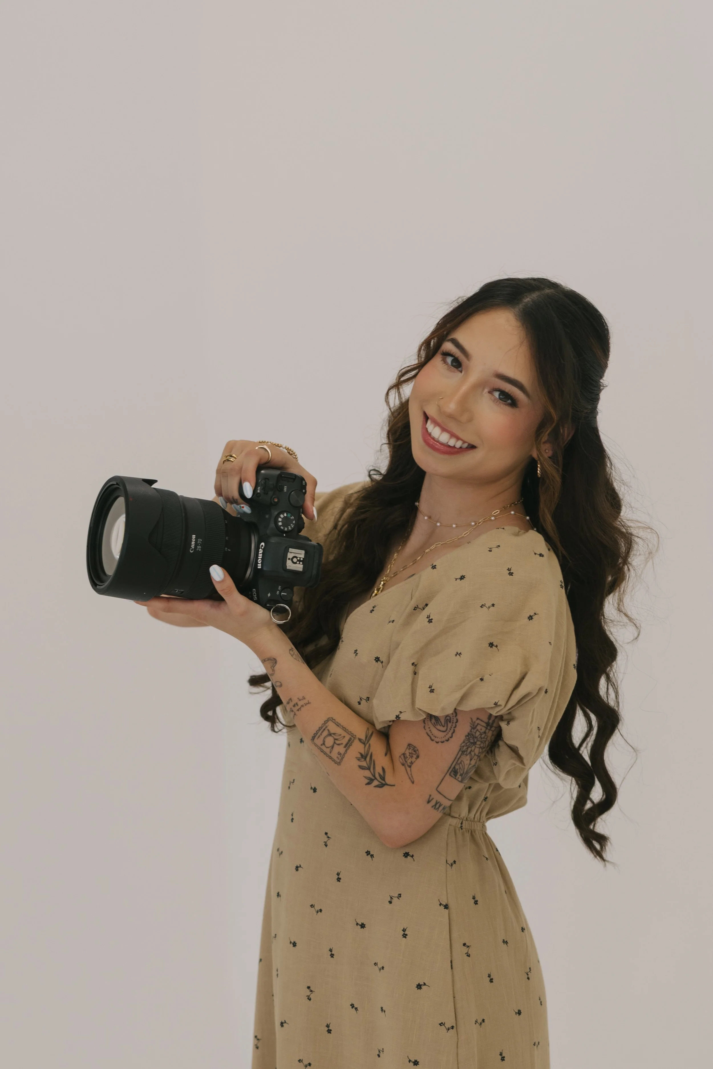 A young woman with long wavy dark hair and tattoos on her arms is smiling while holding a professional Canon camera with a large lens, standing against a plain white background.