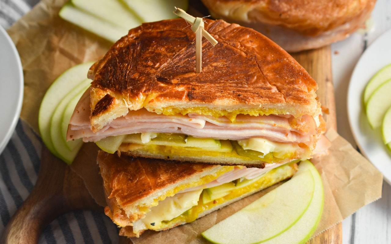Close-up of a sandwich with sliced turkey, cheese, and apple slices on a wooden board.