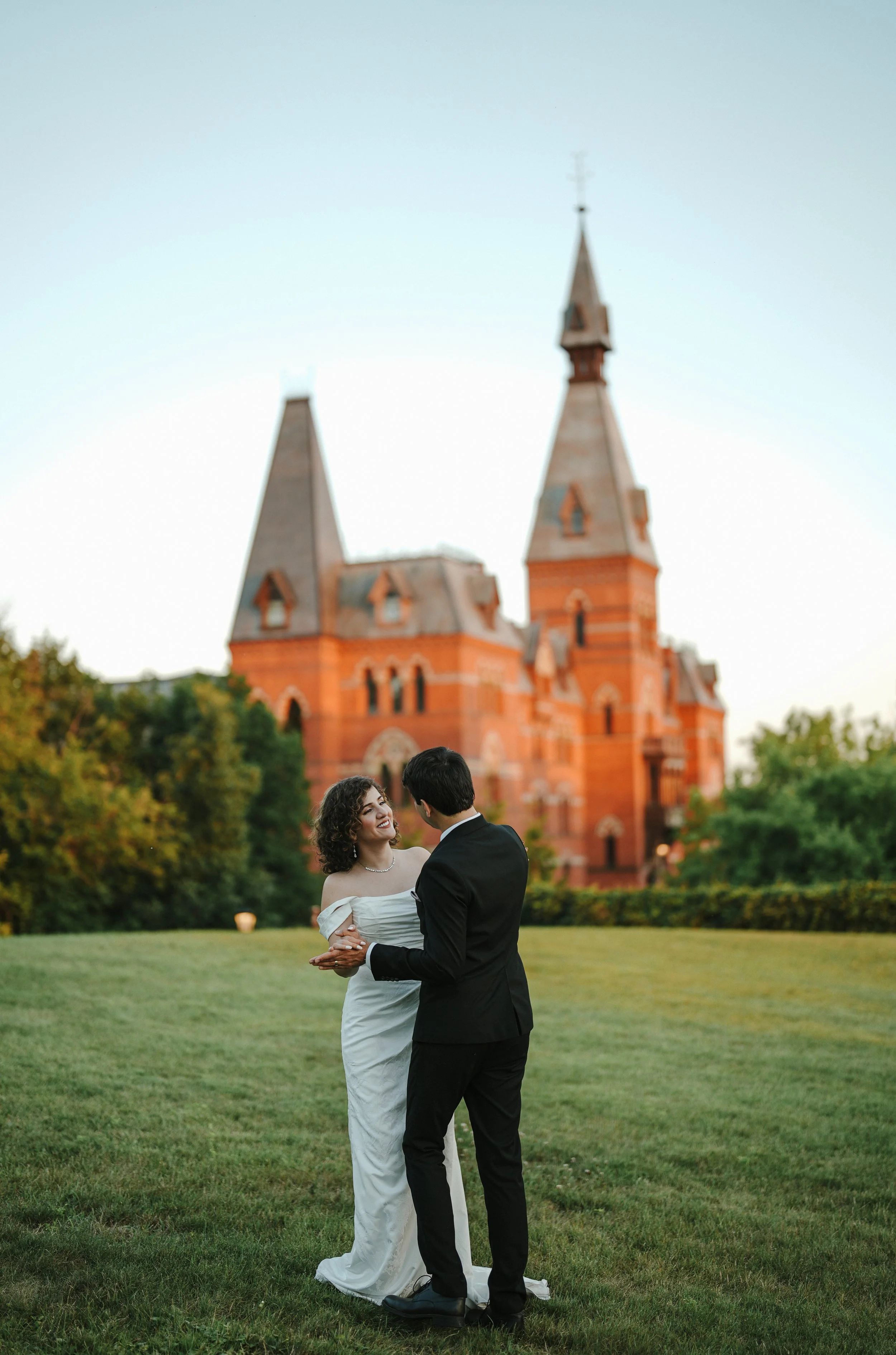 A bride and groom dancing outdoors on a lawn in front of a large red brick church with tall steeples, surrounded by greenery, during sunset.