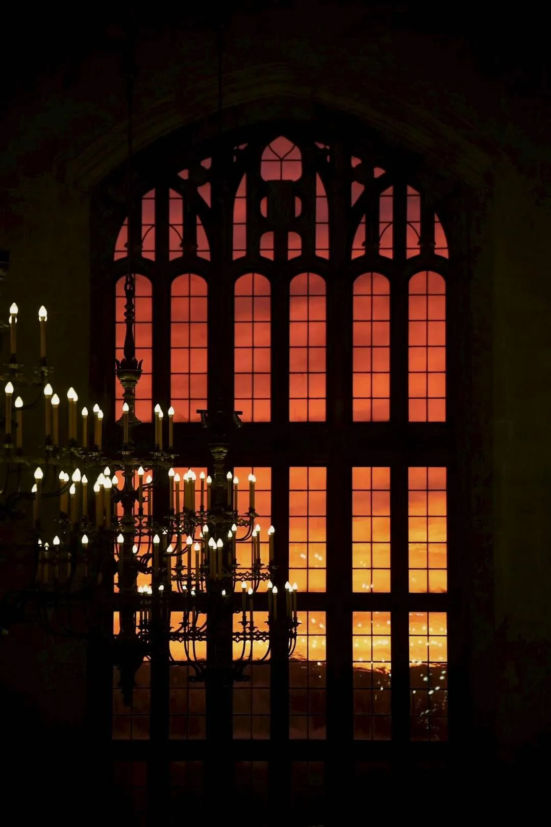A large stained glass window with a sunset view, featuring warm orange and pink hues, and a chandelier with lit candles hanging in front of it.