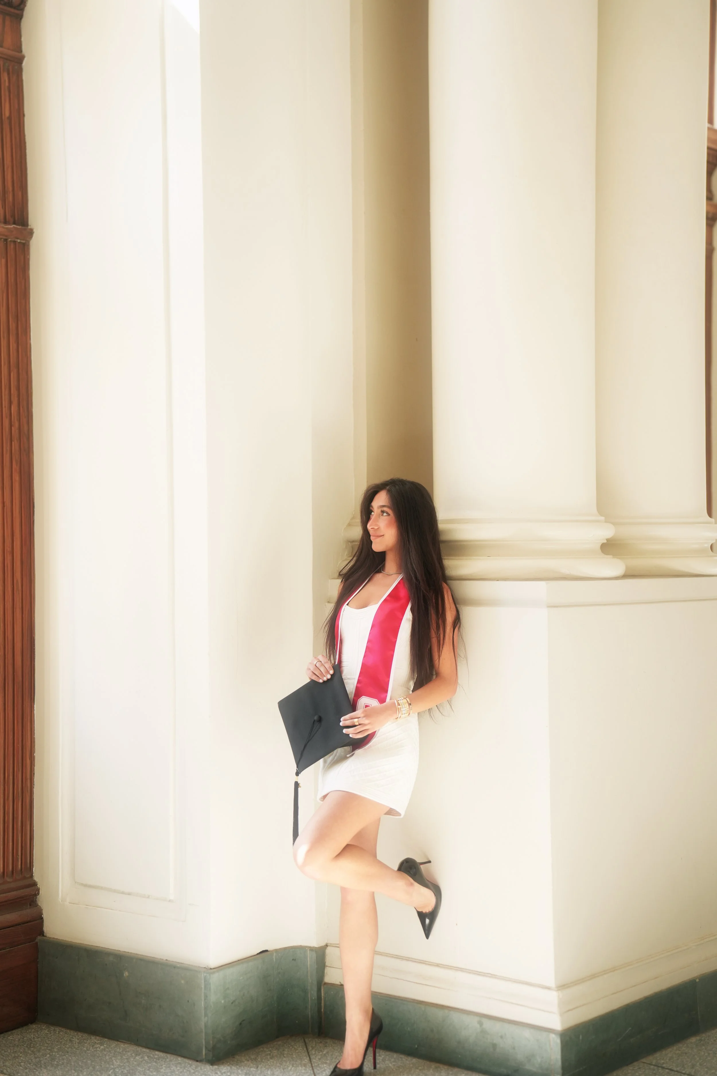 Woman in black heels, white dress, and pink sash, holding graduation cap, standing next to a cream-colored wall with large columns.