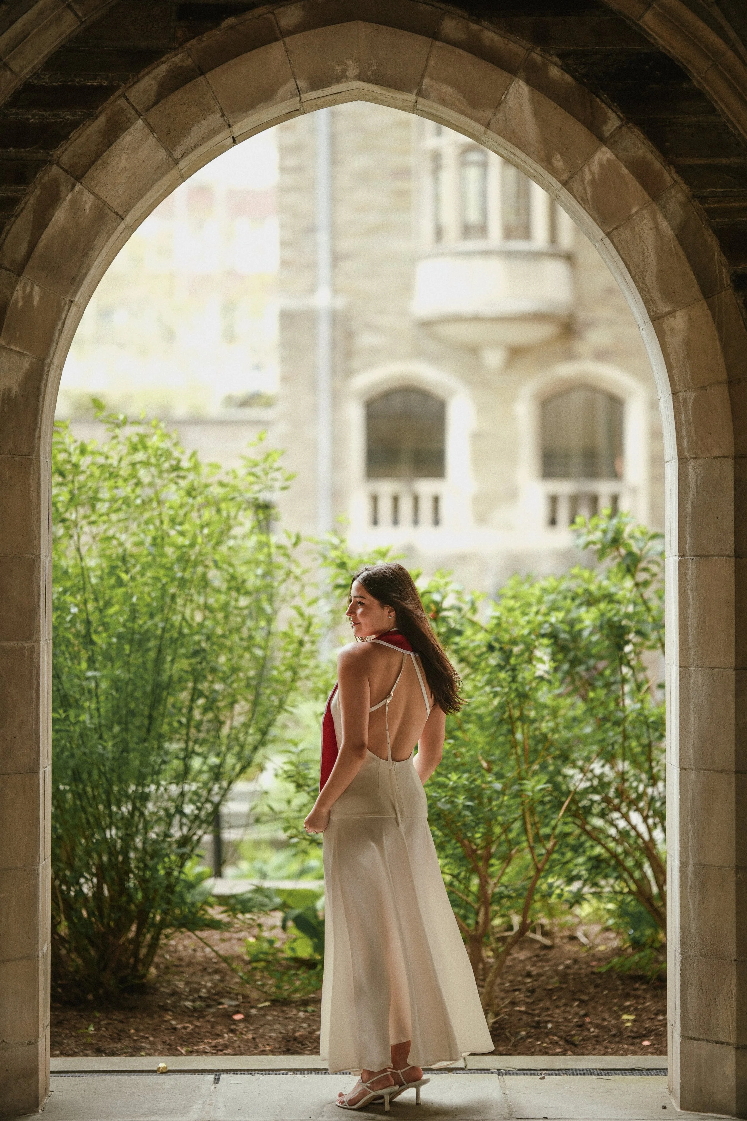 A woman standing under an arched stone doorway, wearing a backless, cream-colored dress and high heels, with greenery and a stone building visible in the background.