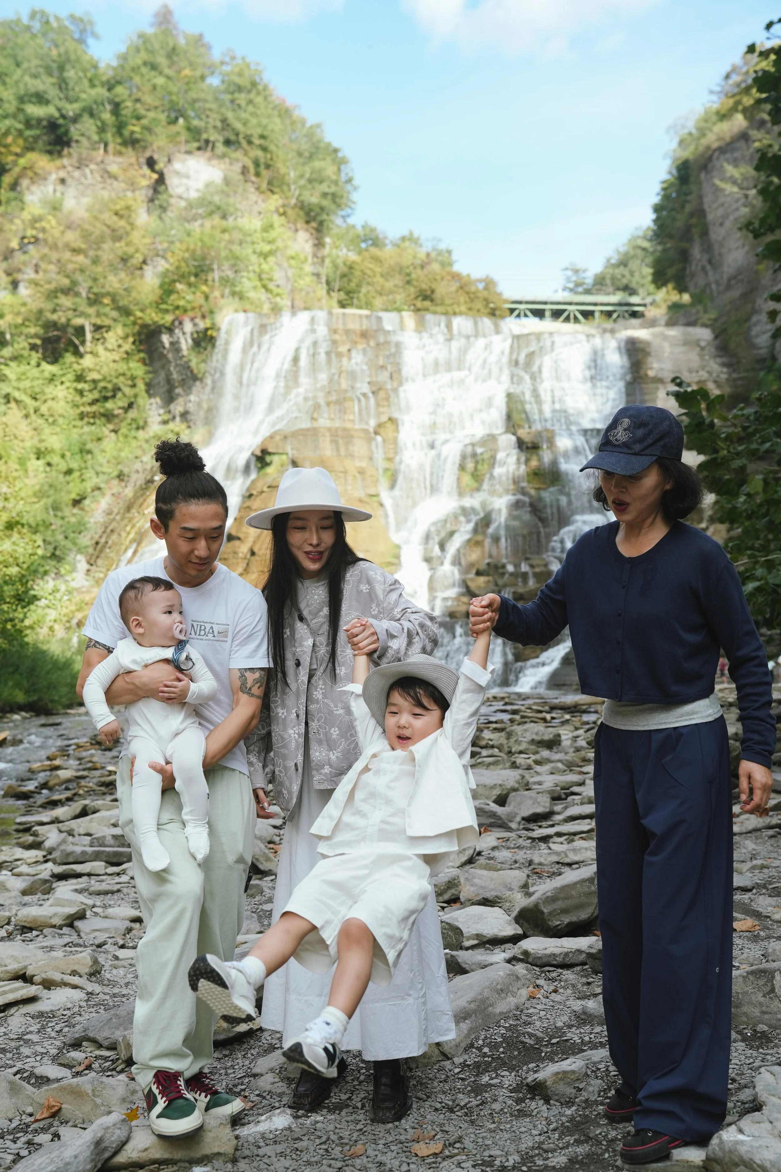 Family of seven, including two young children, standing and walking on a rocky riverbank with a waterfall in the background, surrounded by trees and a partly cloudy sky.