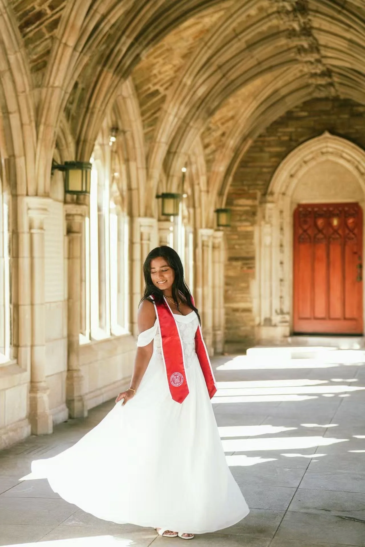 A young woman in a white dress and red graduation stole posing in a stone archway with sunlight streaming in, smiling with eyes closed.