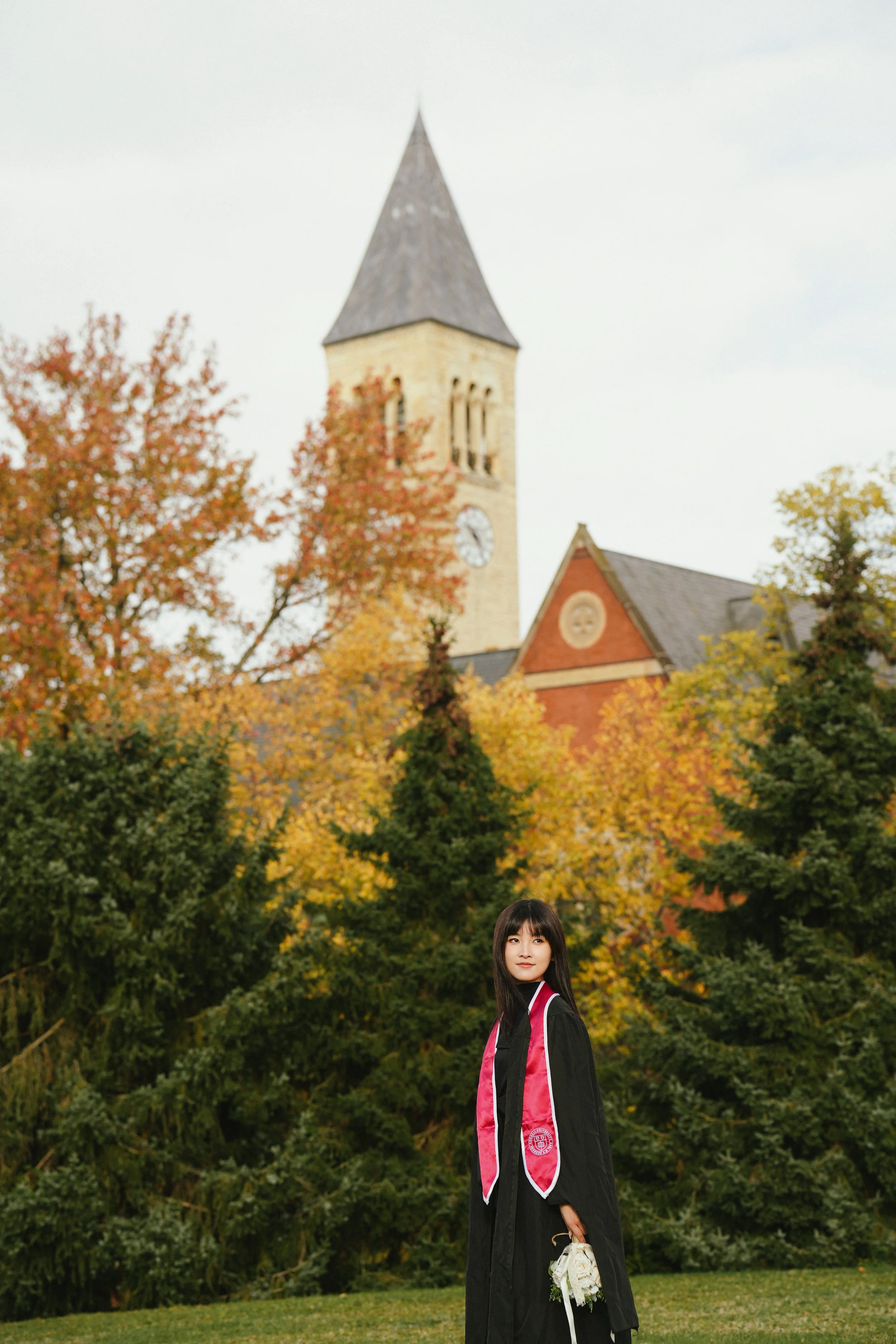 A young woman in graduation attire holding flowers outdoors, with a church and autumn trees in the background.