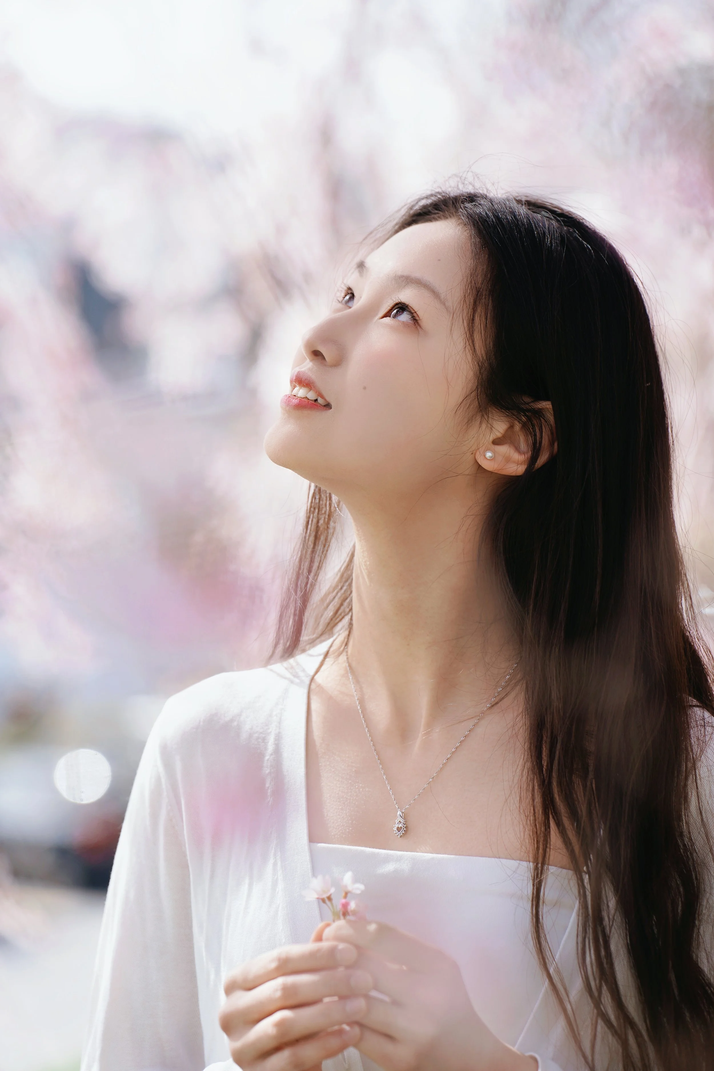 A young woman with long dark hair, looking up with a soft smile, holding pink cherry blossoms in her hands, outdoors with blurred pink and white cherry blossom trees in the background.