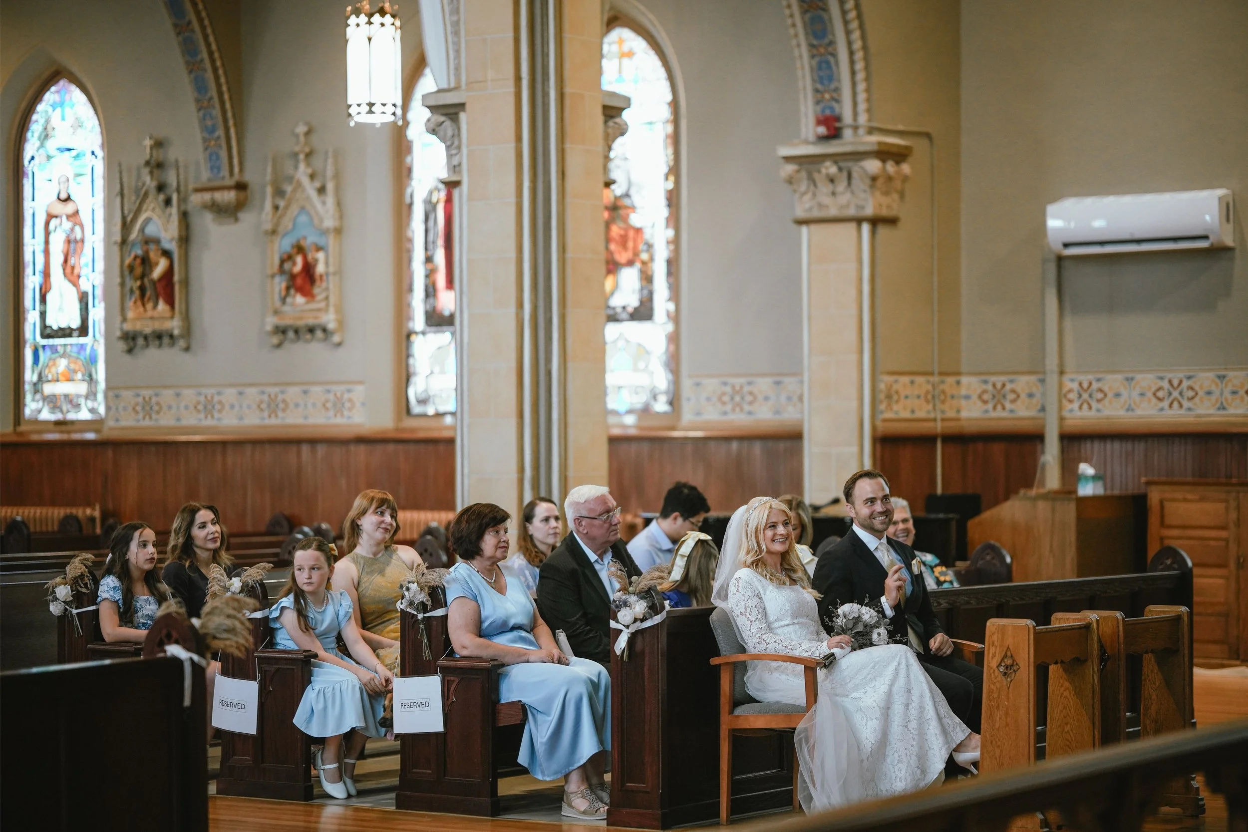 A diverse group of people sitting in a church during a wedding ceremony, with stained glass windows and religious artwork in the background.