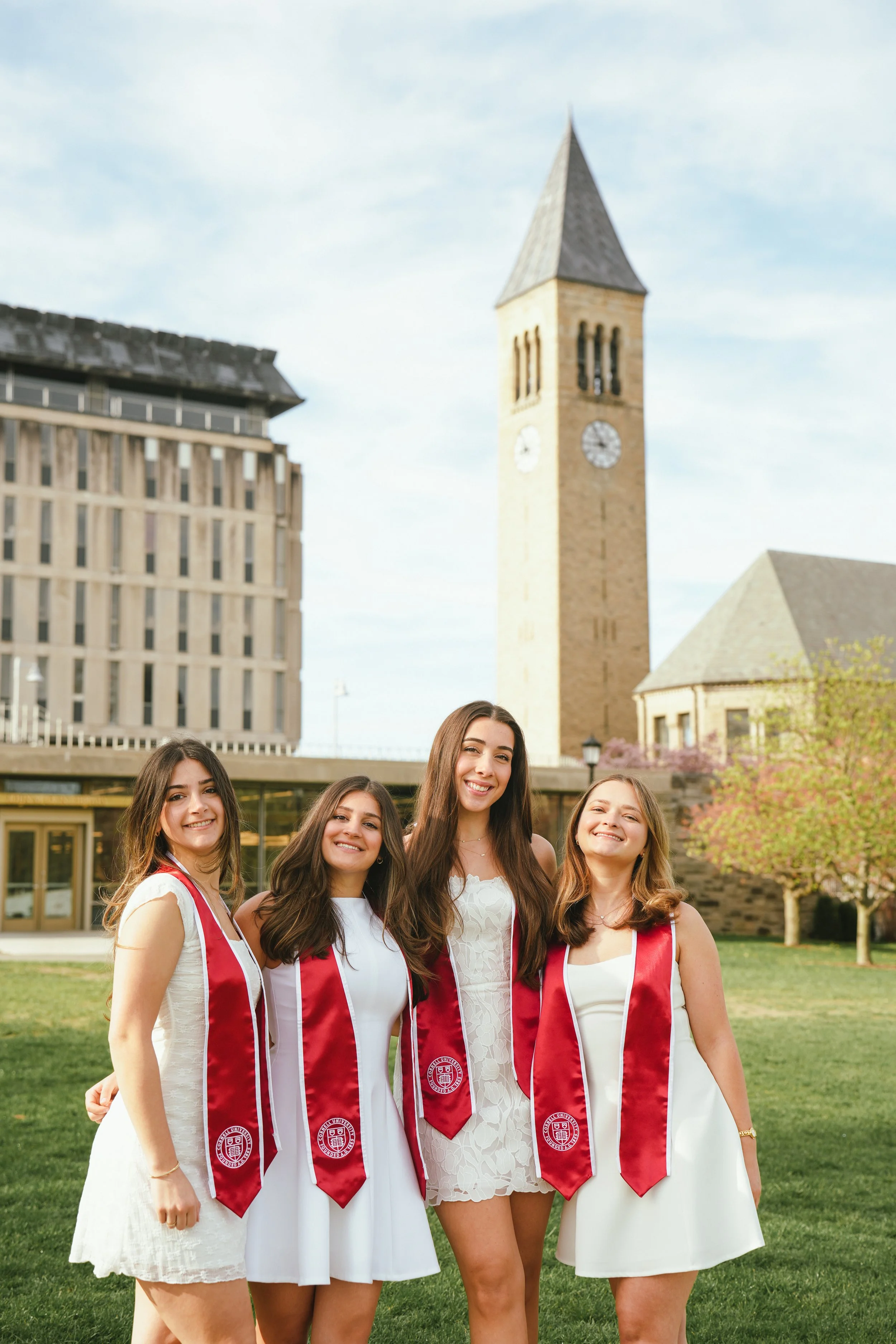 Group of five young women in white dresses and red graduation stoles standing on grass outdoors, with a church tower and a modern building in the background.