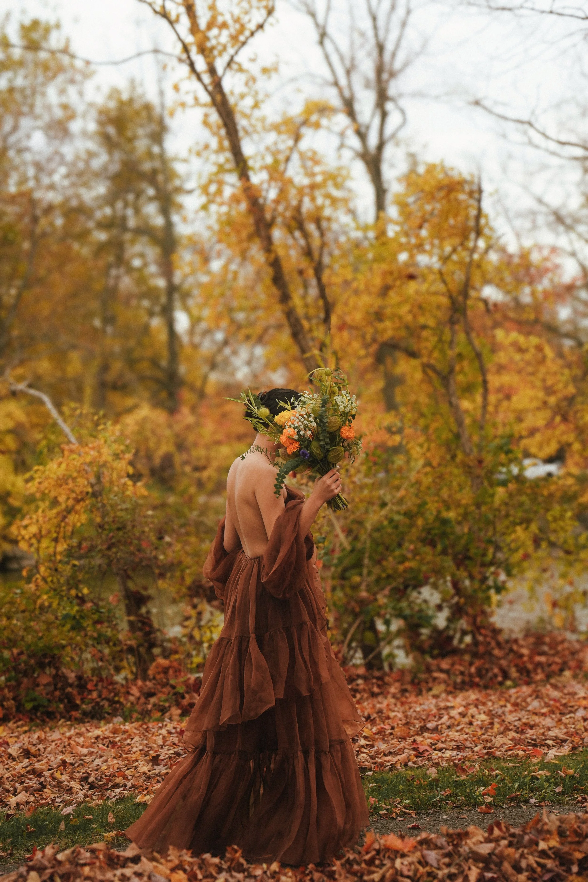 A person dressed in a brown dress holding a large bouquet of flowers covers their face, standing in a park with autumn leaves on the ground and trees with colorful fall foliage in the background.