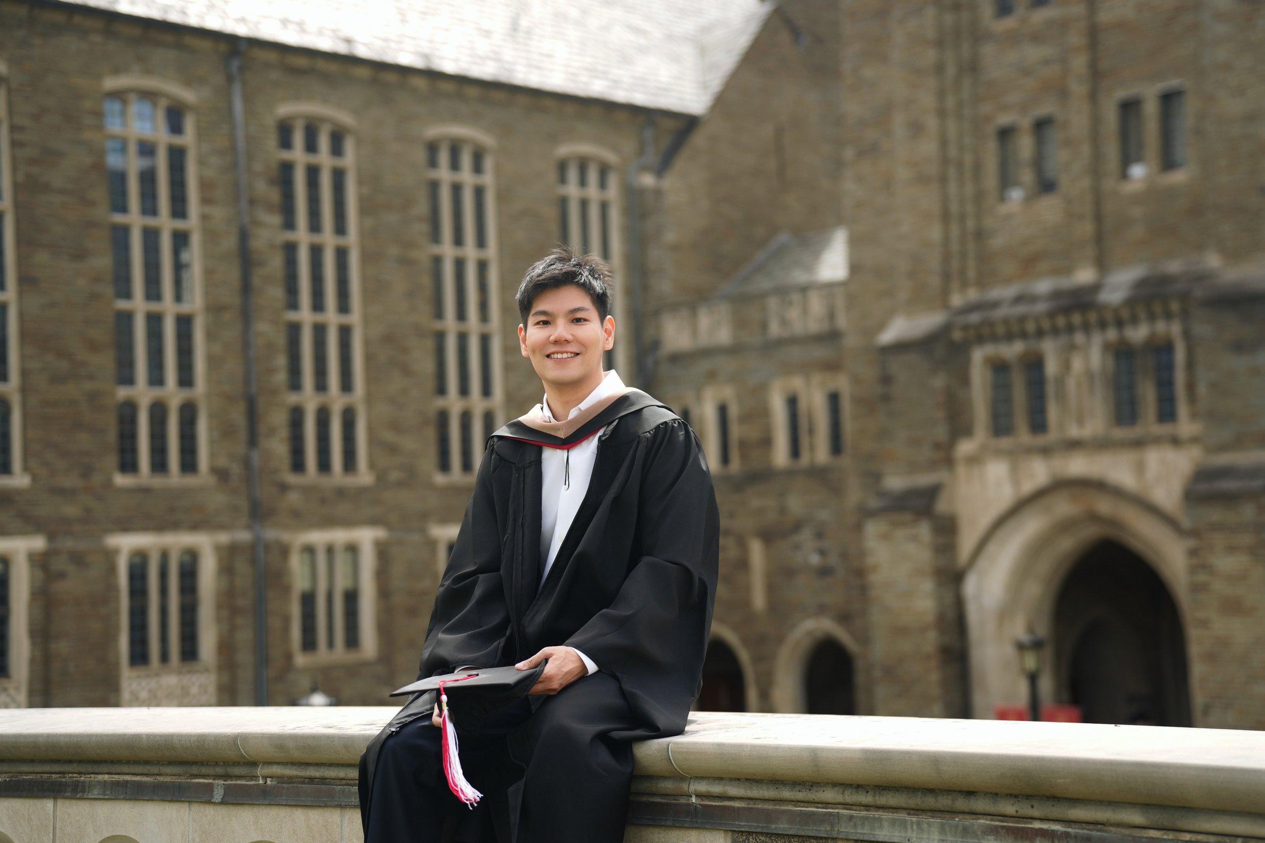 Young man in graduation cap and gown sitting on a stone ledge in front of a historic stone university building.