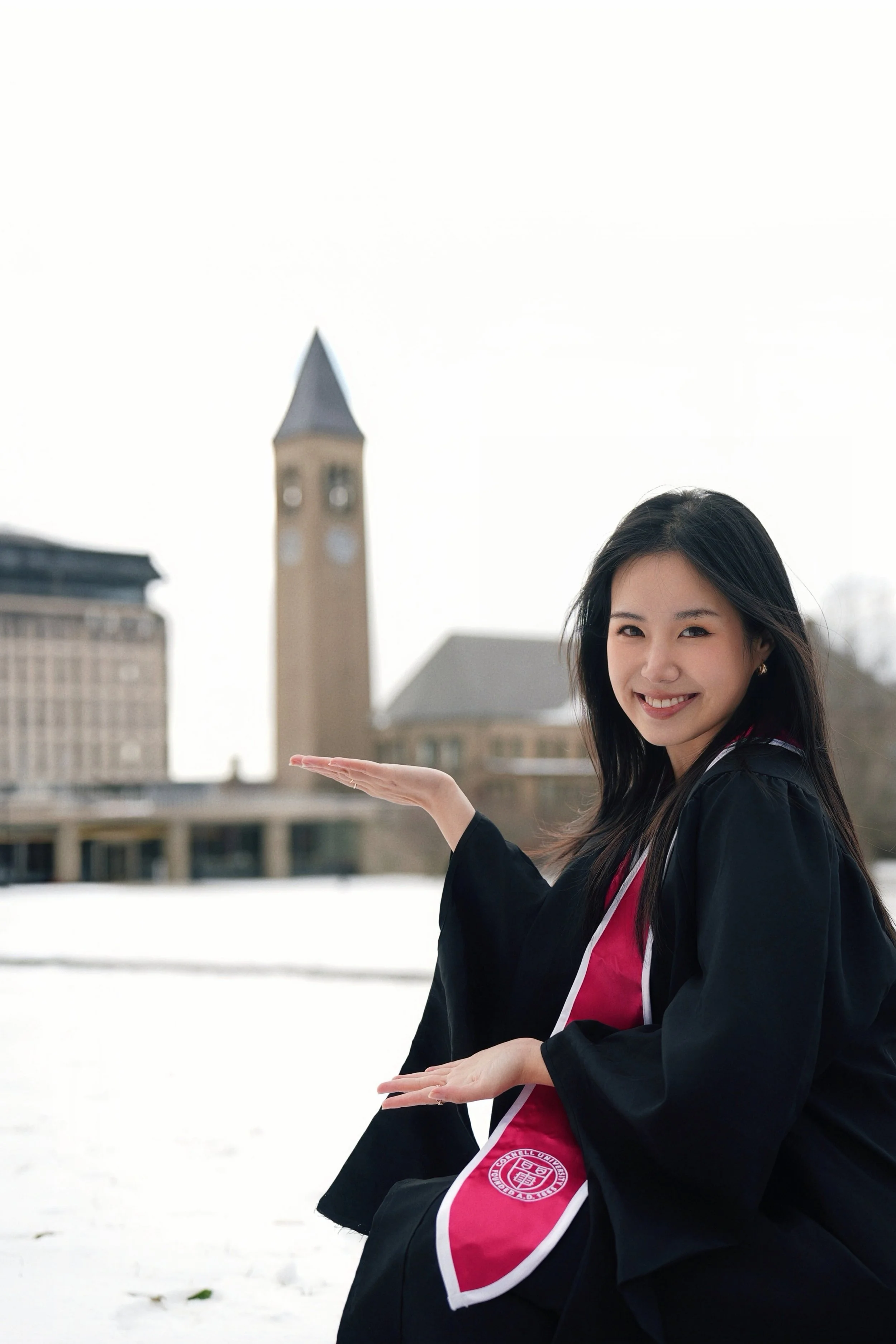 Young woman in a graduation gown smiling and gesturing towards a clock tower and university buildings in the background on a snowy day.